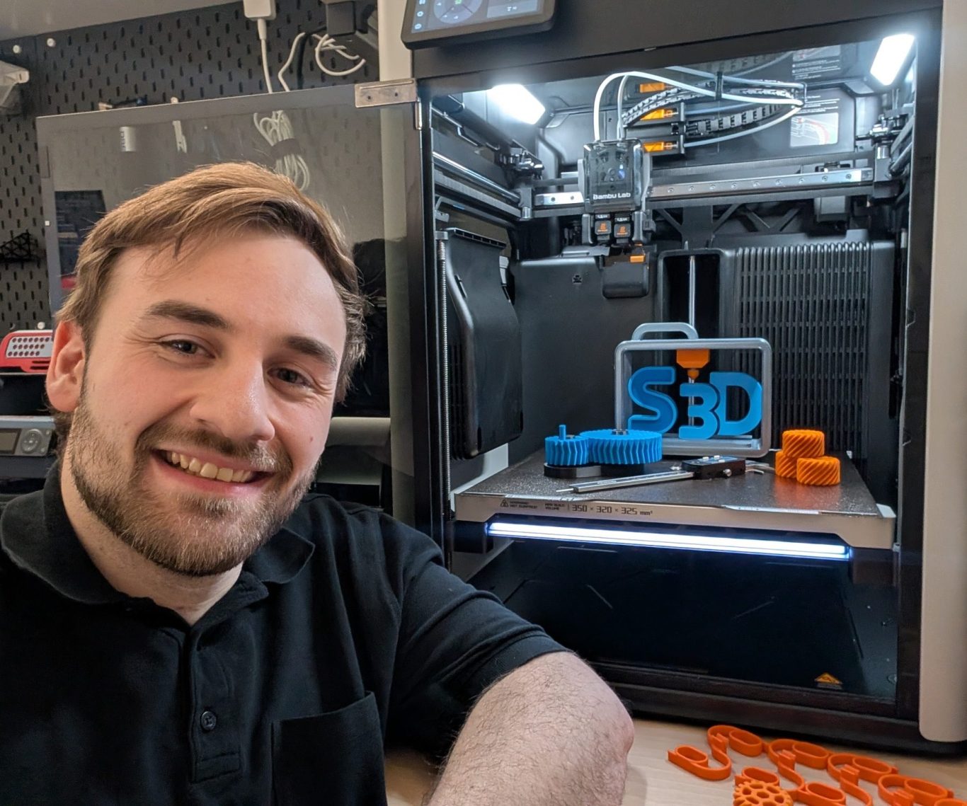 Patrick Schäfer lächelt neben einem 3D-Drucker mit bunten Druckobjekten.